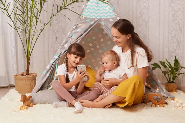 Indoor shot of happy positive family mother with daughters playing at home in wigwam and using smart phone for having fun, watching funny videos or photos.