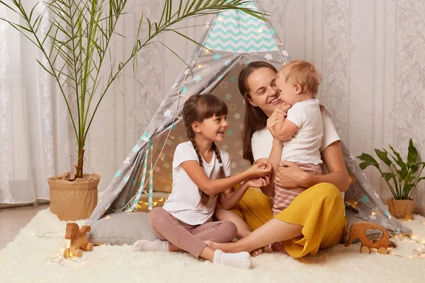 Image of smiling satisfied little girls and her mother playing at home in wigwam and being happy to spend time together, female hugging her kids, being in good mood.