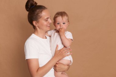Mutlu, koyu renk saçlı bir kadının portresi. Elinde bebek kızıyla duruyor. Anne, kahverengi arka planda izole edilmiş bir şekilde çocuğuna sevgi ve gülümsemeyle bakıyor..