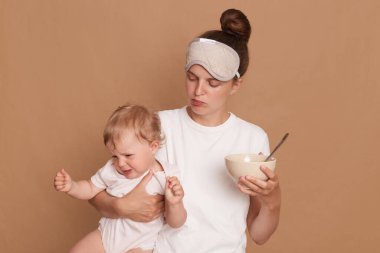Beyaz t-shirt ve uyku maskesi takan bir kadının yatay görüntüsü. Anne, elinde ağlayan bebek kızı ve tabağı tutarken elinde çocuk mamasıyla kahverengi arka planda izole bir şekilde poz veriyor..