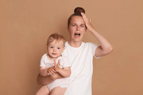 Indoor shot of stressed depressed young mother with bun hairstyle holding her toddler daughter, screaming, being tired and exhausted, touching head, needs rest, standing isolated over brown background