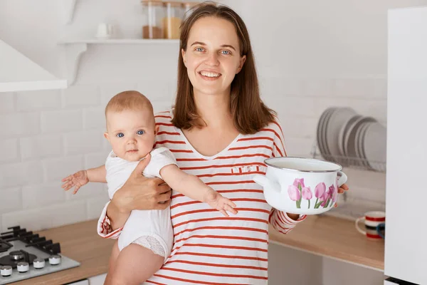 Indoor shot of dark haired woman and small child in the kitchen at home, female in striped shirt holding baby in hands and pot, cooking with her kid, expressing happiness.