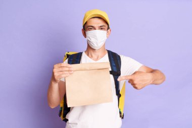 Beyaz tişörtlü, sarı şapkalı ve koruyucu maskeli, kağıt paketli, parmakla karınca parselini gösteren ve emirleri ileten yakışıklı adam kuryesinin kapalı çekimleri..