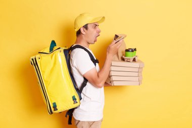 Şaşkın erkek kuryenin şapkalı ve elinde termo çanta ile restorandan pizza kutuları tutarken ve akıllı telefon kullanırken, sarı arka planda izole edilmiş, şok olmuş bir şekilde görüntüye bakarken yan görünüm..