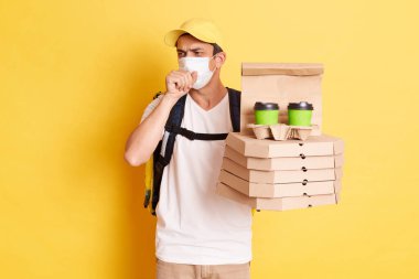 Koruyucu maske takan teslimatçı, elinde pizza kutuları ve paket kahve tutan grip salgını sırasında çalışan kurye soğuk algınlığı belirtileri gösteren, sarı arka planda izole bir şekilde duran bir teslimatçı..