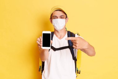 Koruyucu maske takan bir teslimatçının portresi. Elinde termo çanta ve reklam için boş yeri olan telefon ekranını gösteriyor. Sarı arka planda izole edilmiş..