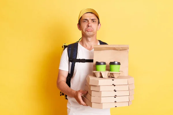Horizontal shot of sad delivery man wearing white t shirt holding food order pizza boxes and coffee, looking at camera with pout lips and expressing sadness, posing isolated over yellow background.