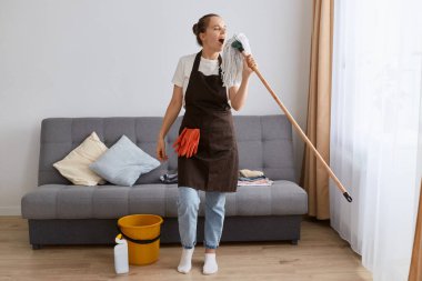 Kapalı alanda saç stili olan çekici bir kadının evde paspas ile yerleri yıkarken, daireyi temizlerken, mikrofon olarak paspas kullanırken, ortalığı temizlerken, şarkı söylerken..