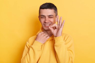 Kapüşonlu komik, pozitif yakışıklı bir adamın yatay görüntüsü. Tamam işareti yapıyor ve kameraya şakayla göz kırpıyor. İyi geri bildirim, sarı arkaplanda izole edilmiş pozlar veriyor..