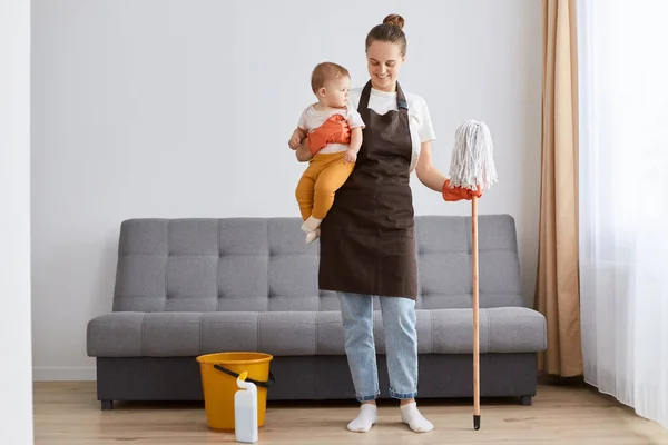 Full length portrait of smiling joyful woman in apron washing floor with mop at home, doing domestic chores, cleaning apartment, holding infant baby in hands.