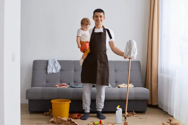 Full length portrait of attractive positive dark haired man in apron doing domestic chores, cleaning apartment together with toddler daughter, expressing positive emotions.