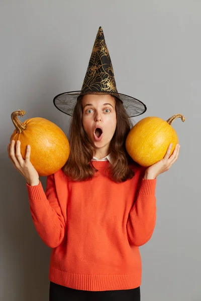 ショックを受けた若い大人の女性の写真魔女の帽子で身を包んだ ハロウィンを祝う 灰色の背景に隔離されたポーズ 手にカボチャとオープン口で立って — ストック写真