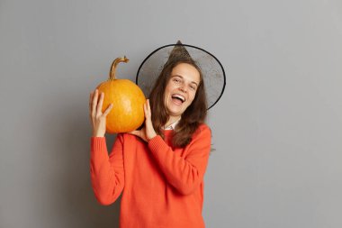 Büyük olgun turuncu balkabağı tutan heyecanlı beyaz kadın, kameraya bakıyor, mutlu bir şekilde gülüyor, Cadılar Bayramı kutlamasında eğleniyor, kazak ve cadı şapkası giyiyor, gri arka planda izole edilmiş..