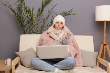 Oturma odasında oturan, kep, eldiven, atkı ve ceket giymiş, elinde akıllı bir telefonla dizlerinin üzerinde dizlerinin üstünde duran, soğuk evde poz veren üzgün, üzgün bir kadının portresi..