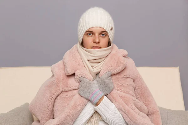 Indoor shot of Caucasian woman wearing cap, gloves, scarf and coat sitting in living room, fells cold, trying to warm up, waiting for start of the heating season.