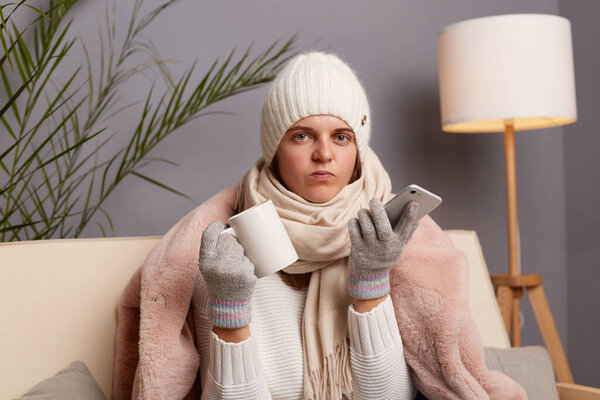 Портрет грустной женщины в шапке, перчатках, шарфе и пальто, сидящей в гостиной с чашкой горячего чая, пытающейся согреться, держащей в руках смартфон, смотрящей в камеру.