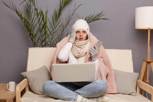 Image of shocked surprised woman wearing cap, gloves, scarf and coat sitting in living room, working on notebook and holding mobile phone, remembering she forgot to pay for central heating.