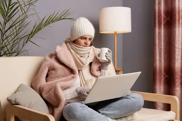 Photo of angry frozen woman wearing cap, gloves, scarf and coat sitting in home interior with cup of hot tea, trying to warm up, working on laptop, looking at display with anger and hate.