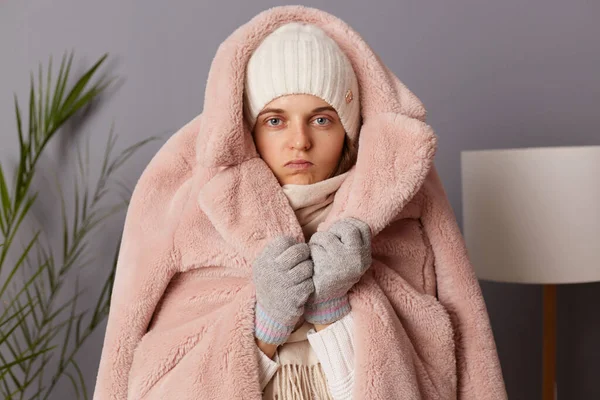 Indoor shot of sad upset Caucasian woman wearing cap, gloves, scarf and coat sitting in living room, puts on warm clothing while being at home in her cold apartment.