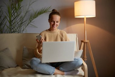 Çekici, beyaz, serbest giyinen, kanepede oturan, dizüstü bilgisayarda çalışan, cep telefonunu elinde tutan, işi için proje yapan bir kadın..