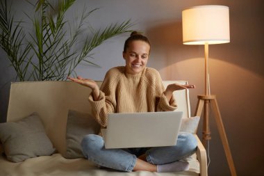 Çaresiz beyaz bir kadının kanepede dizüstü bilgisayarla oturduğu, video görüşmesi yaptığı ya da canlı yayın yaptığı, omuzlarını silktiği, emin olmadığı yatay görüntüsü..