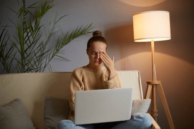 Kapalı alanda, rahat kıyafetler giymiş, kanepeye oturmuş, defter üzerinde çalışırken, gözlerinde acıyı hissederken, gözlerini ovuştururken, uzun süre çalışırken, yorgunluk hissederken,.