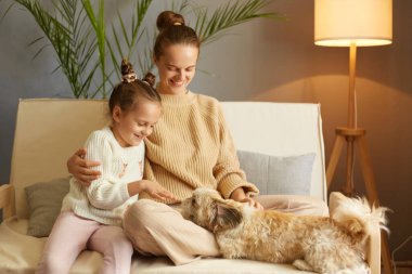 Anne ve sevimli kızın, köpekle oynaştığı, ailenin öksürüğünün üzerinde oturduğu, Pekin köpeğiyle eğlendiği, mutlu bir şekilde gülümsediği yatay bir fotoğraf..