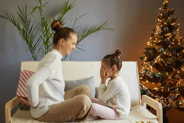 Happy family sharing joy with each other on Christmas. Daughter receives gifts from her loving mother on Xmas Day. Kid covering her eyes, waiting for surprise, sitting in decorated room.