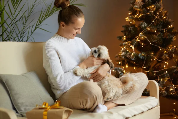 クリスマスツリーの下でギフトやライトで愛らしい犬を愛撫白いセーターのいくつかの笑顔の若い大人の女性 お祝いの部屋で犬と幸せな若い女性 ハッピー ホリデー — ストック写真