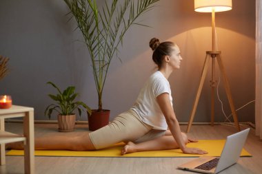 Beyaz tişörtlü, sıska atletik bir kadının portresi. Rahat odada yoga yapıyor. İnternette ders izlemek için dizüstü bilgisayar kullanıyor..
