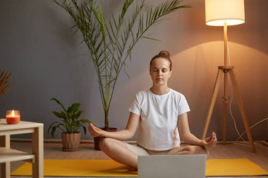 Kapalı mekanda, beyaz tişörtlü, rahat ve sakin bir kadının rahat bir çekimde rahat bir odada yoga yaparken, nilüfer minderinde otururken, Mudra jesti ve kapalı gözlerle..