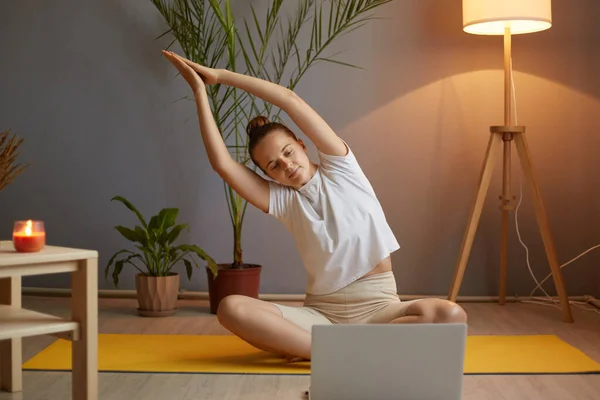 Full length portrait of attractive woman with hair bun wearing white t shirt practicing online yoga at home in cozy room, looking at laptop display, raised hands in praying gesture, stretching.