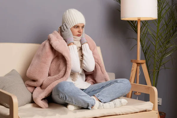 暖かい服を着て 部屋のソファに座ってスカーフで包まれた悲しい不幸な女性の屋内ショットは 彼女の耳を暖めようとし 中央暖房をオフにして家の中でポーズ — ストック写真