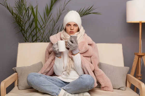 Image of sad upset Caucasian woman wearing warm clothes and wrapped in scarf sitting on sofa in room with cup of hot tea, holding mobile phone, looking at camera.