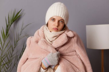 Oturma odasında kep ve palto giyen, depresyonda olan, soğuk bir dairede ısınamayan üzgün bir kadının kapalı mekândaki görüntüsü..
