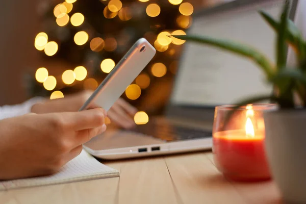 Side view of female person\'s hand holding mobile phone in hands, unknown woman using cell phone, browsing internet with blurred Christmas tree on background.