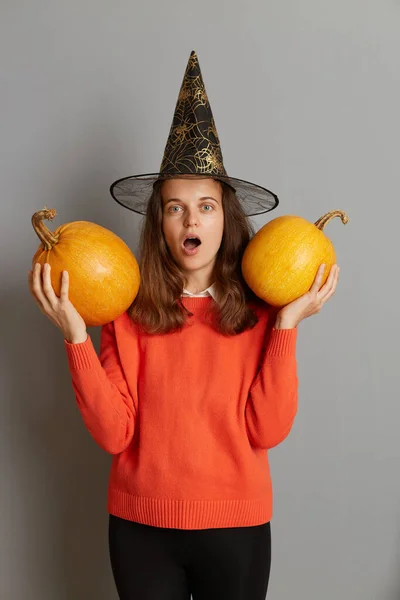 Portrait of surprised little scared Caucasian girl standing with pumpkins in her hands,wearing witch hat posing with Halloween pumpkin standing on gray background, looking at camera with open mouth.
