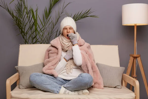 Full length portrait of sleepy woman posing in cold room sitting on the cough wearing warm hat, mittens and cap, needs to have nap, yawning, covering mouth with hand.