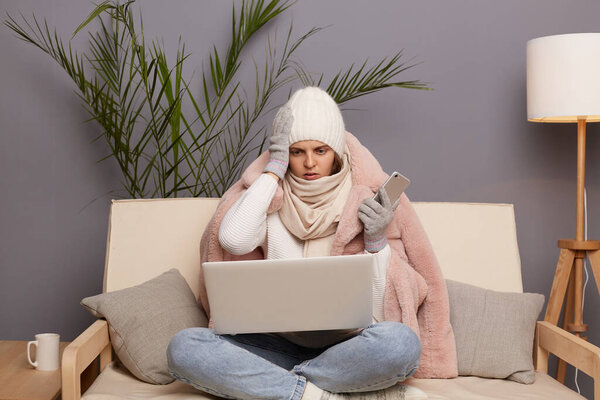 Portrait of shocked young adult woman posing in cold room sitting on the cough wearing warm hat, mittens and cap, working online on laptop, having problems with her work.