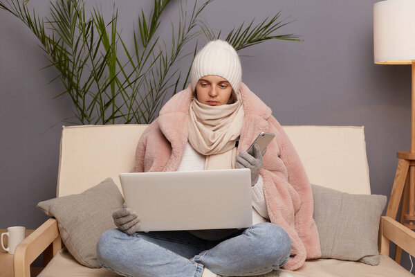 Horizontal shot of sad unhappy Caucasian woman wearing cap, gloves, scarf and coat sitting on sofa with laptop and smart phone in hands, being busy, posing in cold room.