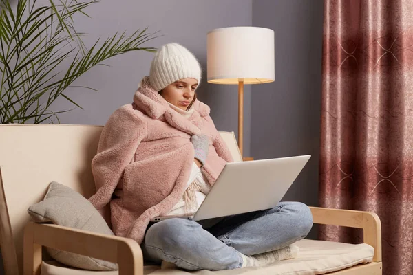 Side view of attractive frozen woman being in cold room sitting on the cough wearing warm hat, mittens and cap, getting warm in fur coat while working online at home.