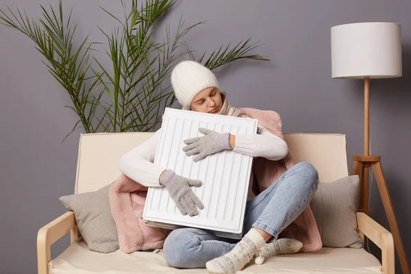 Indoor shot of sleepy frozen woman in coat and hat sit in cold living room and embracing radiator, feels freeze at home, having nap while getting warm, keeps eyes closed.