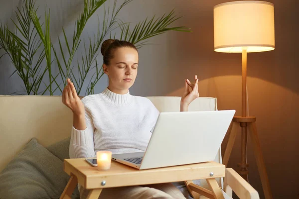 Indoor shot of calm concentrated woman wearing white sweater sitting on sofa and trying to relax while working on laptop, practicing yoga at work.