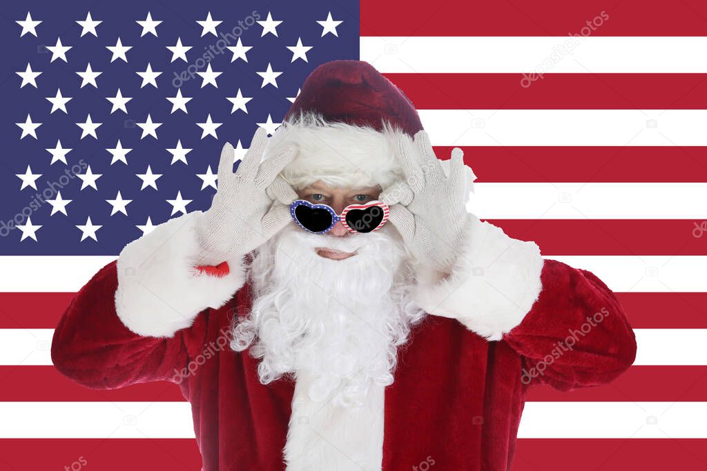Santa Claus smiles as he puts on his Sunglasses with an American Flag Background. Merry Christmas and Happy Holidays. Merry Christmas And Happy New Year. Greeting Card With Santa Claus Sitting in front of the American Flag. Winter Holidays. Ho Ho Ho.