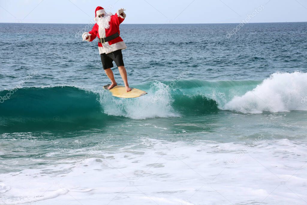 Surfing Santa. Santa Claus is riding a surf board. Santa Claus in sunglasses surfing in the Ocean. Santa Claus Surfs on his Surf Board while on a Beautiful Beach with a Blue Ocean. Christmas Vacation.