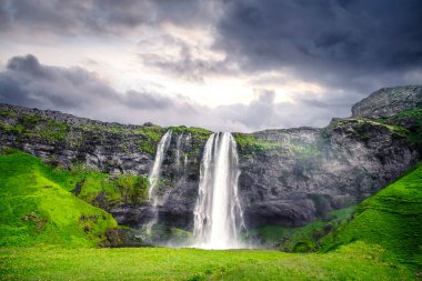 İzlanda 'daki güzel Seljalandsfoss şelalesi lav kayalıkları ve bulutlu bir günbatımında yeşil tarlalarla çevrili.