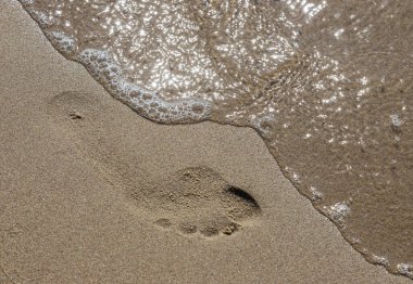 Deniz kenarında yalınayak yürürken sarı kumsalda ayak izlerini silip süpüren bir adamın ayak izleri. Hayatın tefekkürü