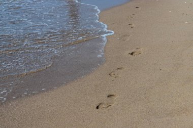 Deniz kenarında yalınayak yürürken sarı kumsalda ayak izlerini silip süpüren bir adamın ayak izleri. Hayatın tefekkürü