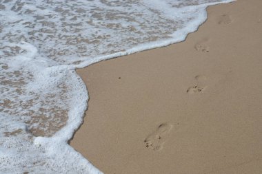 Kumsalda ayak izleri. Kumdaki ayak izleri bir deniz dalgasına karşı. Güneşli bir günde altın kum, sahil, dalga ve ayak izleri..