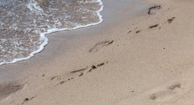 Deniz kenarında yalınayak yürürken sarı kumsalda ayak izlerini silip süpüren bir adamın ayak izleri. Hayatın tefekkürü